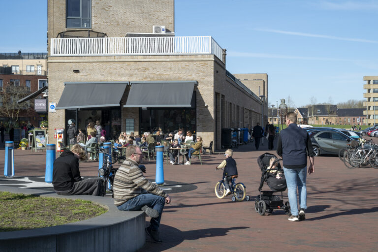 Foto van de wijk Schuytgraaf in Arnhem. Je ziet mensen zitten op een bankje, een vader achter een kinderwagen en een terras waar mensen wat zitten te drinken.
